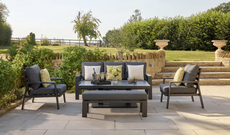 Grey Bramblecrest premium garden furniture set: sofa, chairs, table with flowers on patio amid greenery.