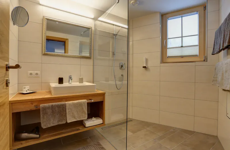 Modern hotel bathroom with wooden vanity, white tiles, glass shower, towels, and window.