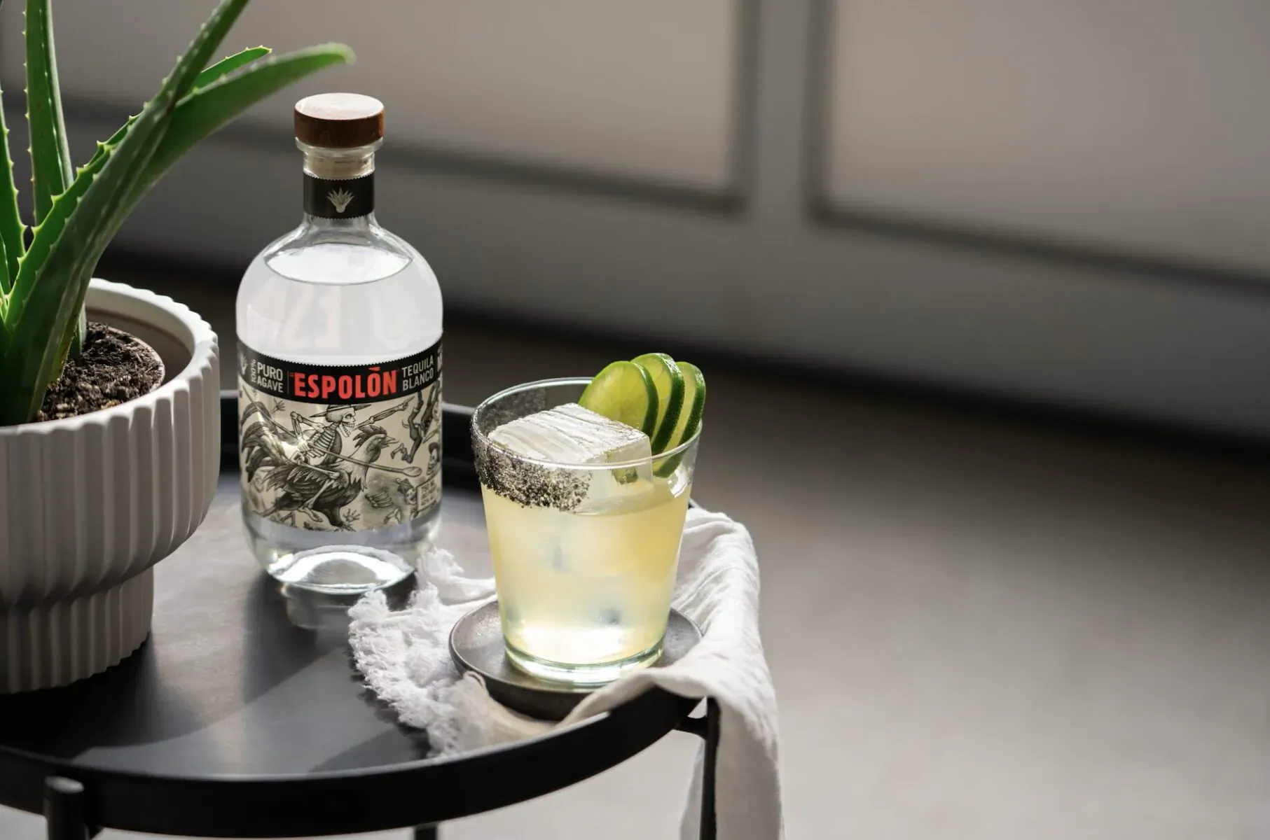 Espolón Tequila bottle beside a margarita garnished with lime wedges and salt rim on a table with aloe plant.