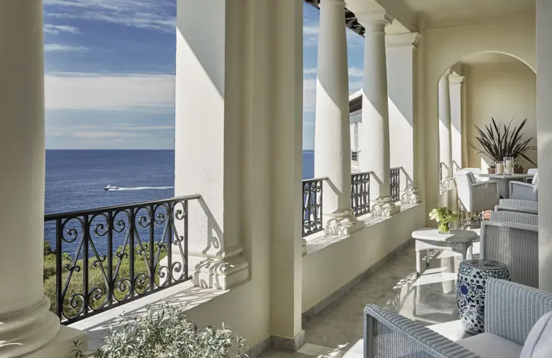 Elegant balcony with white columns overlooking blue ocean, wicker furniture, plants, and balustrade.
