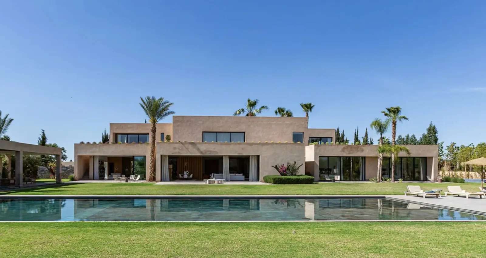 Luxury beige villa with large glass walls, palm trees, pool, and lush lawn in sunny Marrakech setting