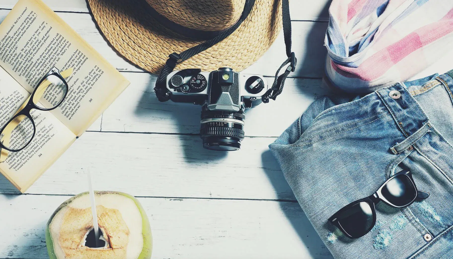 Flat lay of beach essentials: straw hat, vintage camera, sunglasses, open book, jeans, coconut drink on white wood.