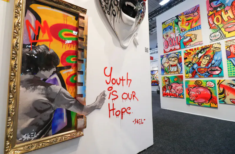 Framed black-and-white photo of man painting 'Youth is our hope - MZ' in red graffiti on white wall amid colorful art gallery at Artexpo Dallas.