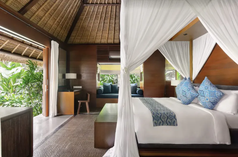 Luxurious Balinese villa bedroom with thatched roof, white canopy bed with blue pillows, and tropical forest views.