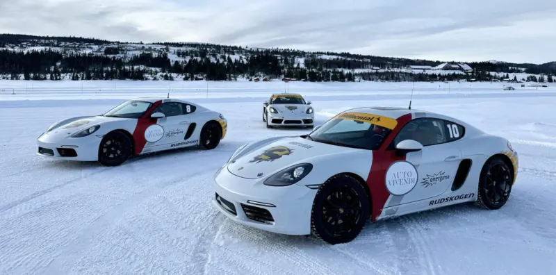 Three white Porsche sports cars with red accents and circular logos drifting on a snowy frozen lake amid winter mountains.