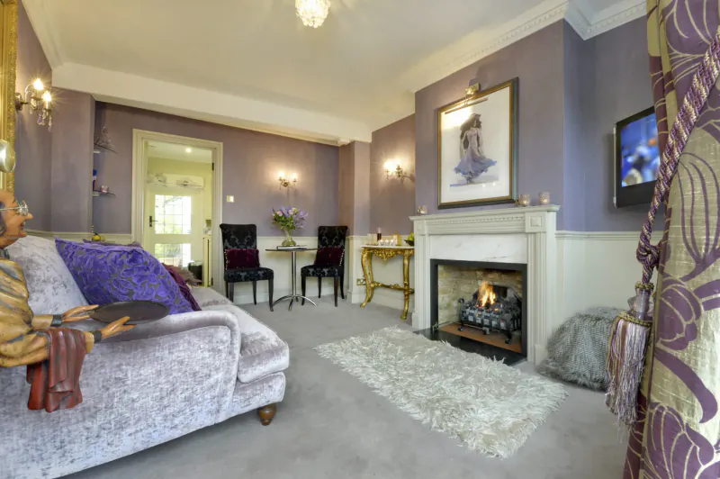 Luxurious living room in Bray Cottage with purple velvet sofa, lit fireplace, gilt chairs, floral artwork, and heavy drapes.