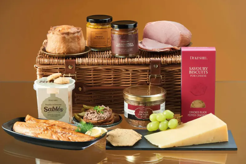 Wicker hamper overflowing with Dukeshill hamper biscuits, Sabbi mix, ham, cheese, pâté, smoked salmon, jars, and grapes.