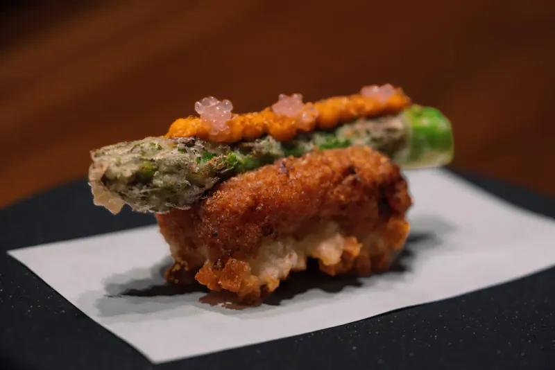 Asparagus tempura sushi topped with orange urchin roe and pink flowers on white paper at UMU.