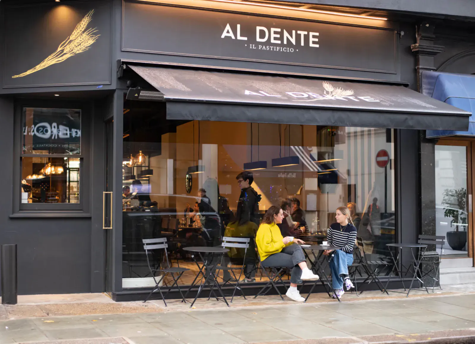 Al Dente pasta shop exterior with golden wing logo, awning, large windows showing interior, patrons at outdoor tables on city street.