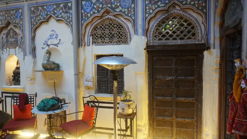 Ornate Rajasthan haveli courtyard with blue intricate arches, red chairs, heater, and traditional decor lit warmly.