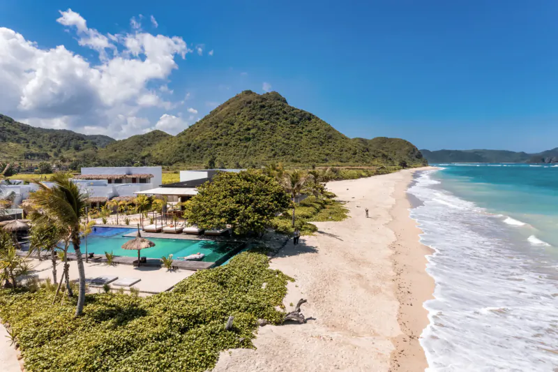 Aerial view of Amber Lombok Beach Resort with infinity pool, white sand beach, turquoise sea, and lush green mountains under blue sky.