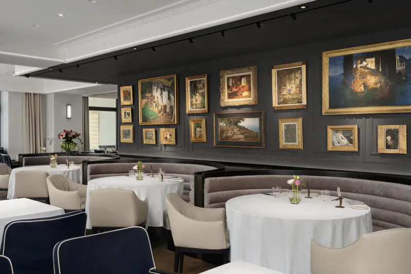 Elegant Vienna gallery restaurant with ornate framed paintings on dark walls, round tables, upholstered chairs, and fresh flowers.
