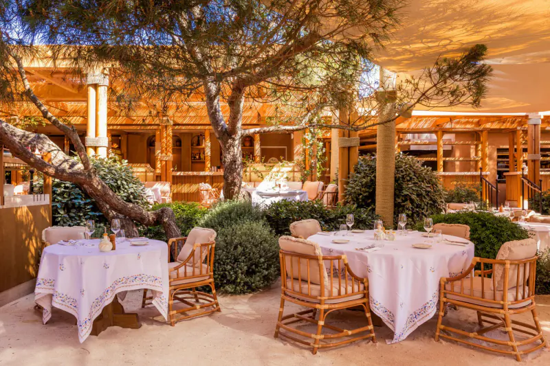 Elegant outdoor dining area at luxurious beach house with white-clothed tables, wicker chairs, trees, and wooden architecture under shade.
