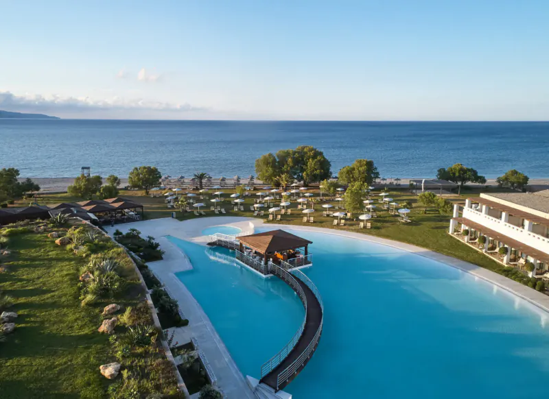Aerial view of Giannoulis Cavo Spada Crete resort: infinity pool with wooden bar, sunbeds, lush gardens, and sea beyond.