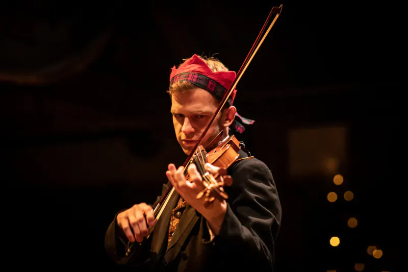 Violinist with red headband playing violin on dimly lit Christmas stage with bokeh lights.