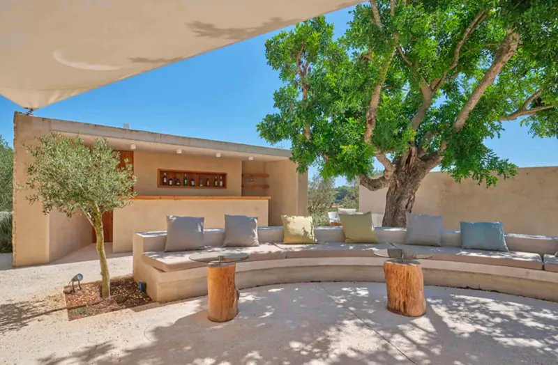 Outdoor seating area at Son Penya hotel with curved beige bench, cushions, tree stumps, olive tree, and large green tree under blue sky.