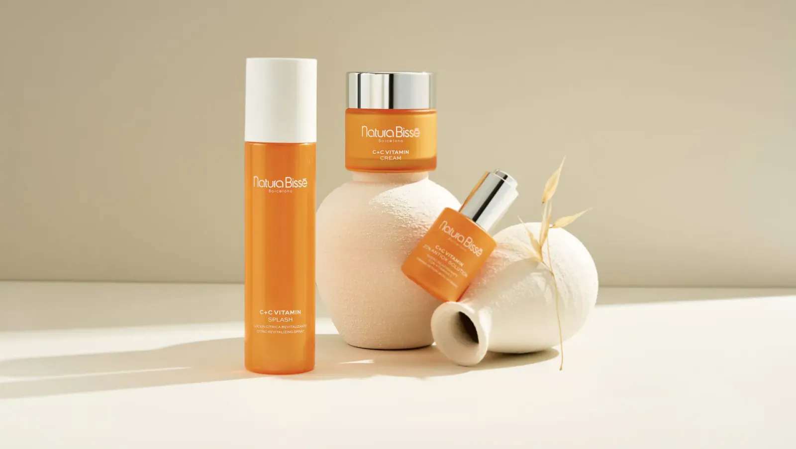 Natura Bissé sun protection products: orange skincare bottles and jars on white surface with dried grass.