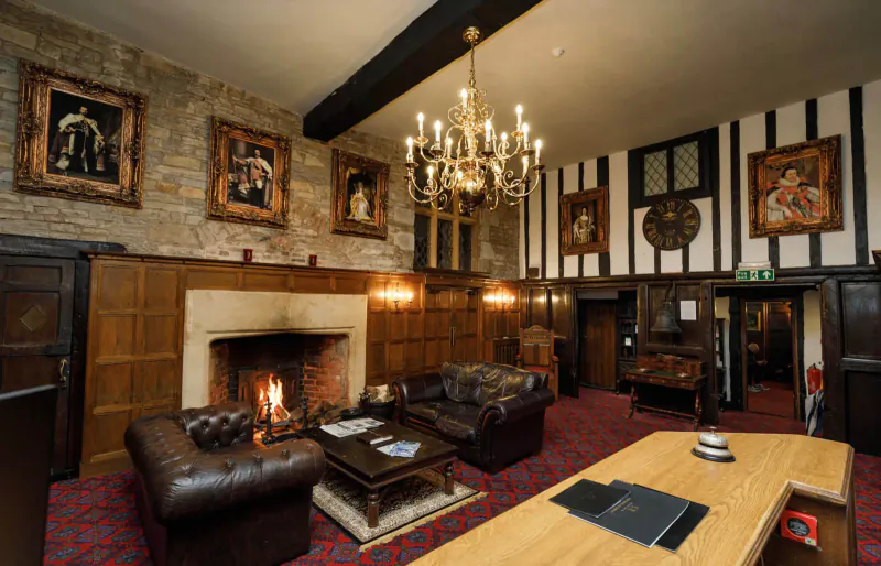 Elegant Tudor lounge at Karma Salford Hall with lit fireplace, leather sofas, chandelier, and portraits on wood-paneled walls.
