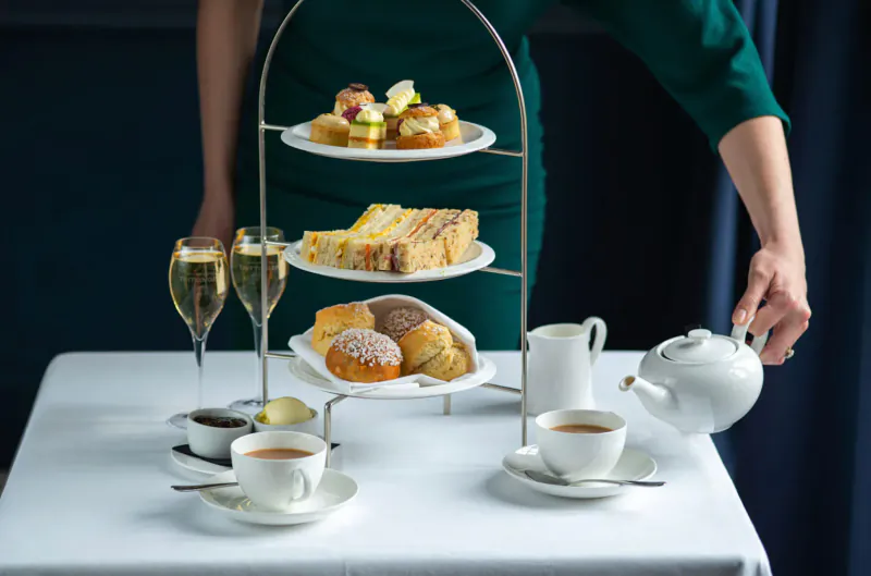 Woman in green dress serving tiered stand of sandwiches, scones, cakes, tea, and champagne at elegant table, Royal Crescent Hotel.