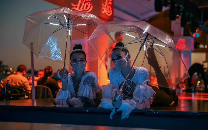 Two performers in frilly white outfits and sunglasses hold clear umbrellas on Lio Cabaret stage, Ibiza.