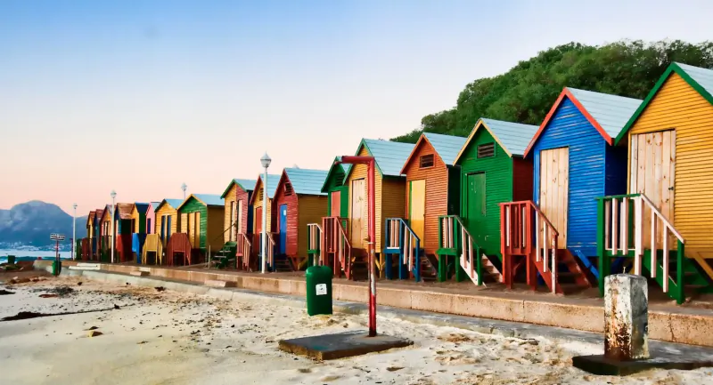 Row of colorful beach huts in vibrant blues, greens, yellows along Kalk Bay shore at sunset, Cape Town