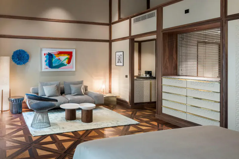 Elegant hotel suite with wooden floors, gray sofa, coffee tables, colorful abstract art, and king bed.