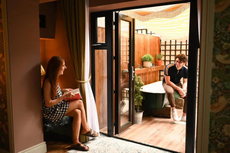 Woman reading book seated by open doors to balcony with man relaxing near potted plants and tub at quirky hotel retreat