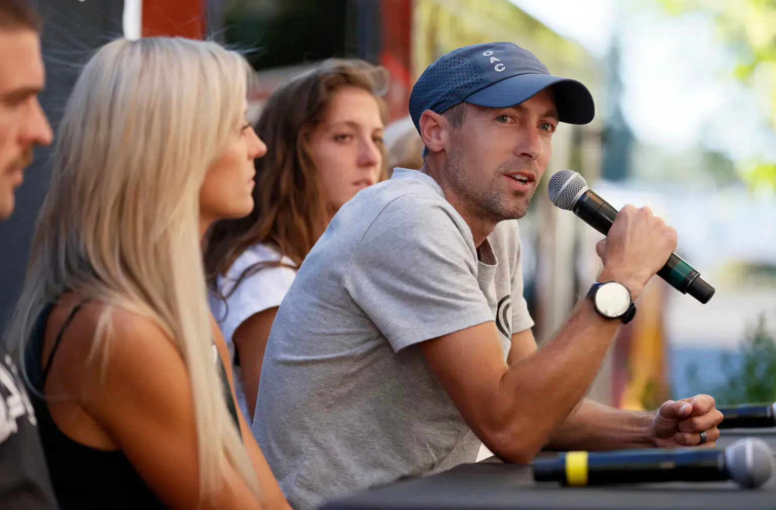 Man in blue cap speaking into microphone at panel with two blonde women, 'Run for each other' event.