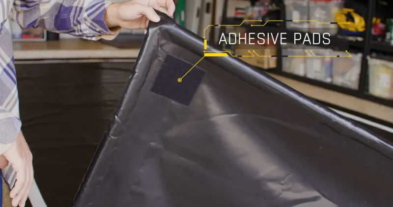 Person holding black GaraMat garage floor mat with yellow 'ADHESIVE PADS' label on black pad, in garage setting.