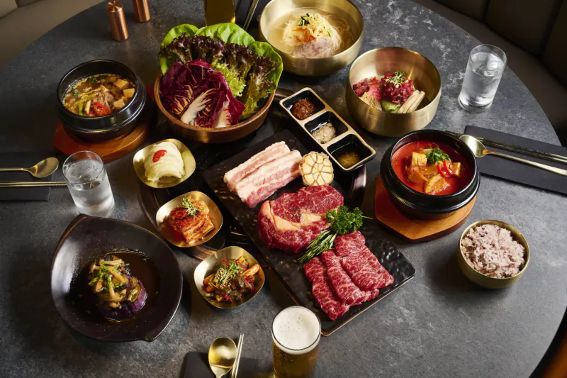 Korean BBQ spread on table: grilled pork belly and beef, kimchi, stews, banchan, rice, beer
