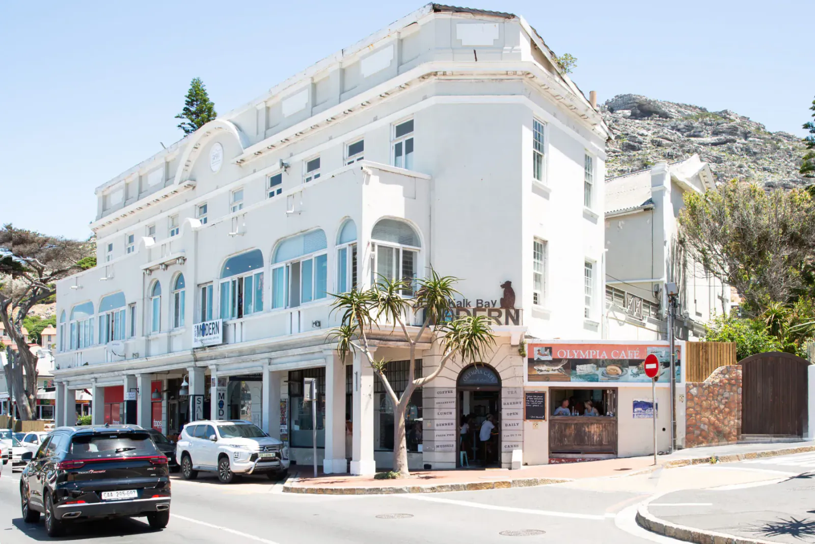 White corner Olympia Café building in Kalk Bay, with blue windows, balcony plants, cars on street, and mountain backdrop.