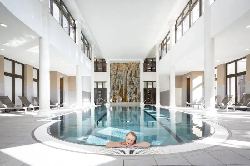 Young red-haired girl relaxing in a circular indoor pool in a luxurious white spa with stone wall art and lounge chairs.