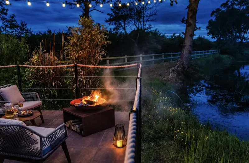 Cozy evening deck at The Boathouse lodge with fire pit, cushioned chairs, lanterns, string lights, overlooking river.