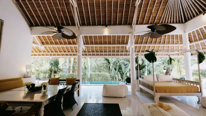 Luxurious open-air Bali villa interior with wooden ceilings, ceiling fans, dining table, lounge, and infinity pool.