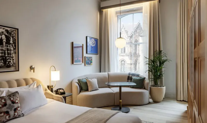 Cozy Virgin Hotel room in Edinburgh with beige sofa, round table, plants, art, and large window.