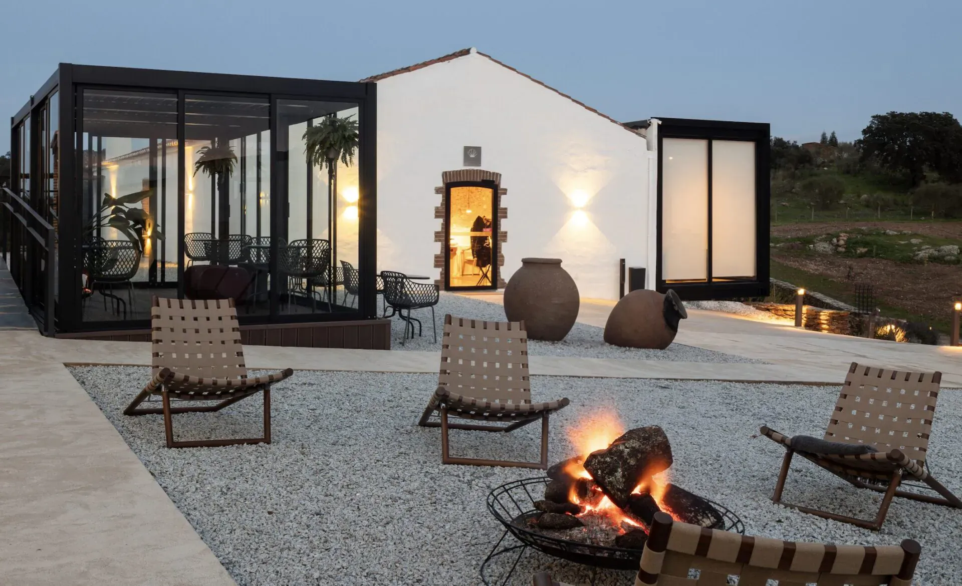 Modern white house with glass extension at dusk, outdoor gravel patio with woven chairs around a fire pit, large terracotta pots, plants, and rural landscape.