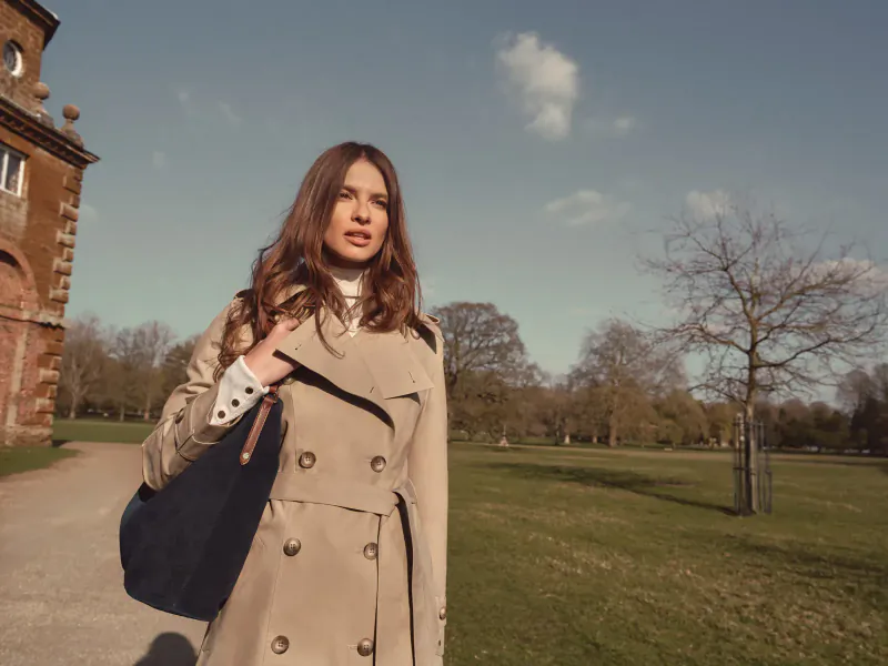 Woman in beige trench coat and white collar, black bag, walks path by brick manor house in sunny park with bare trees.