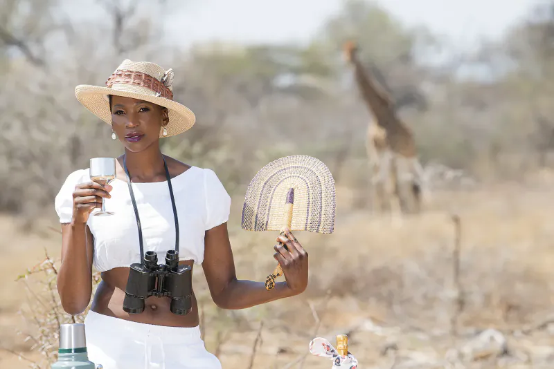 Black woman in white outfit holds wine glass and fan, binoculars around neck, wide-brim hat, giraffe in South African savanna safari background