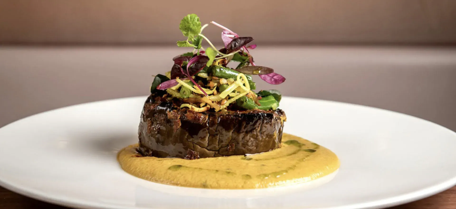 Seared eggplant steak on yellow sauce, topped with purple and green herbs and sprouts, on white plate.