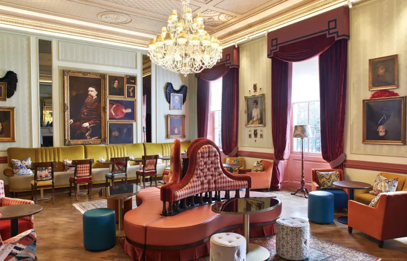 Elegant Home House gallery lounge with chandelier, ornate portraits, velvet curtains, and colorful armchairs.