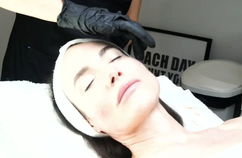 Dr Kubicka in black gloves performs facial treatment on relaxed woman with head wrap on bed, 'Each Day' sign behind in clinic.