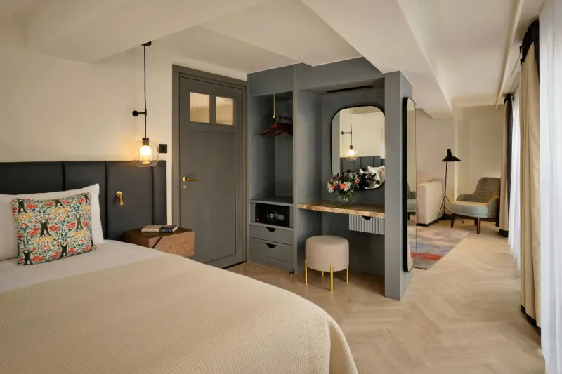 Modern canalside hotel room with king bed, gray vanity with flowers, armchair, and city-view drapes.