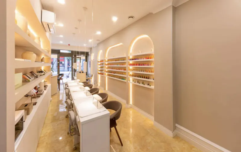 Modern nail salon interior with white manicure stations, chairs, shelves of nail polishes, and arched doorways in chic Fitzrovia spa.