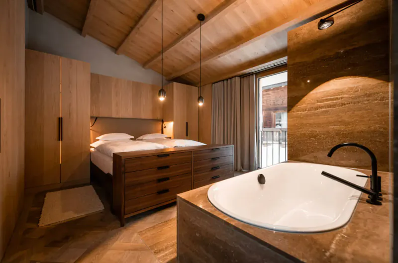Luxurious wooden Austrian chalet bedroom with king bed, freestanding bathtub, balcony view.