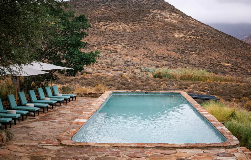 Infinity pool with teal lounge chairs on stone deck at Simbavati Cederberg Ridge retreat, rugged mountains backdrop