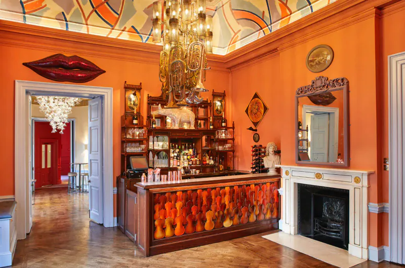 Elegant Home House club bar with orange walls, art deco ceiling, large lips artwork, chandelier, and fireplace.