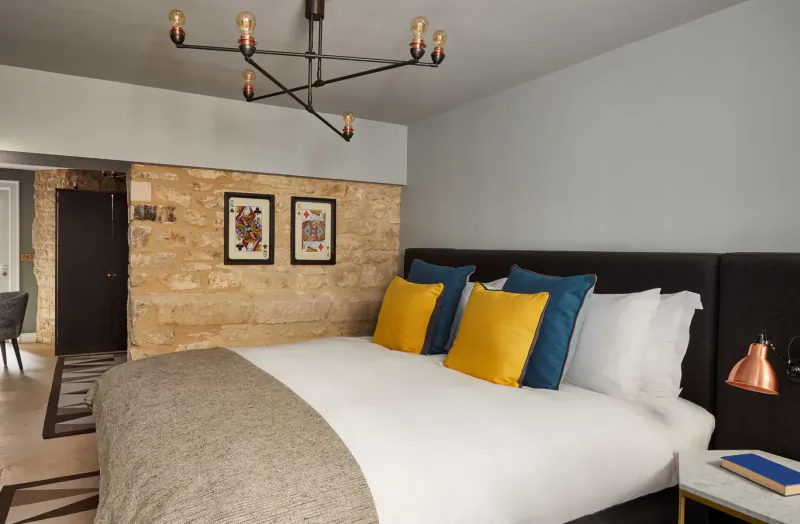 Cozy Hotel Indigo Bath bedroom with black headboard, yellow and teal pillows, throw blanket, chandelier, art on stone wall, bedside lamp.