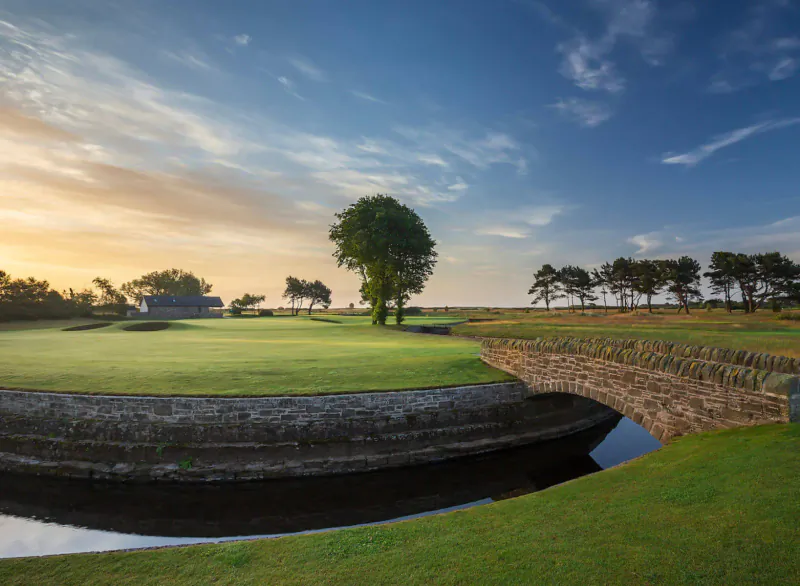 Carnoustie Golf Links: green with stone bridge over stream, trees, clubhouse at sunset