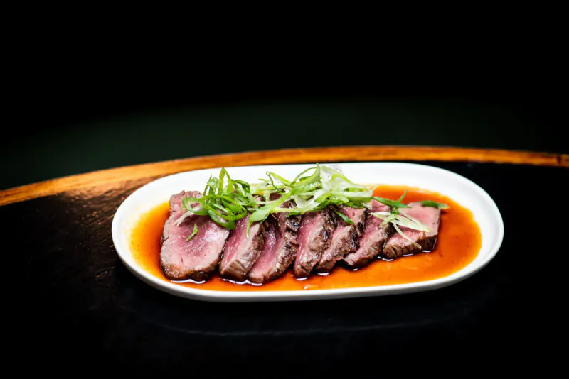 Six slices of seared pink beef on orange sauce in white dish, garnished with green onions, on wooden table.