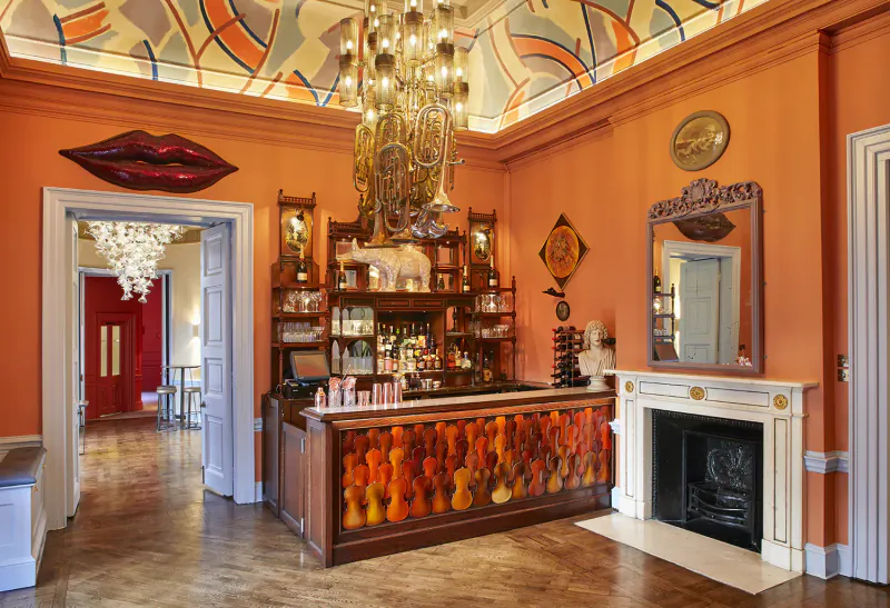 Elegant Home House bar with orange violin bar front, chandelier, art deco ceiling, large lips artwork, and fireplace.
